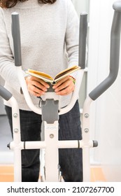 Unrecognized woman exercising on an elliptical bike while reading a book. Concept of multitask, doing sport at home.