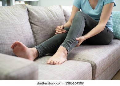 unrecognized young asian woman had pain at calves sitting on couch at home. chinese female in jeans hands massage legs on grey sofa. day off people sick illness painful body indoors in apartment. - Shutterstock ID 1386164309