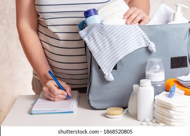 Unrecognizible Pregnant Caucasian Woman In Striped T-shirt Packing Big Blue Diaper Bag To Maternity Hospital. Diapers, Nappy, Hat, Bottle And Other Necessary Things For Newborn Baby.