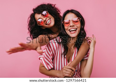 Unrestrained merriment of two girls captured on snapshot. Photos in pink shades of brunettes with beautiful curls, embracing in friendly way
