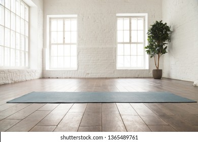 Unrolled yoga mat on wooden floor in modern fitness center or at home with big windows and white brick walls, comfortable space for doing sport exercises, meditating, yoga equipment