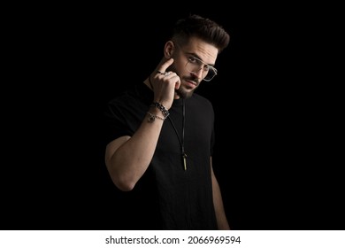 unshaved cool young man with beard and glasses touching face with finger and posing on black background in studio