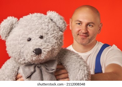 An Unshaven Bald Man With A White T-shirt With A Blue Strap Gives A Gray Teddy Bear With A Bow On A Red Background. A Holiday Gift.