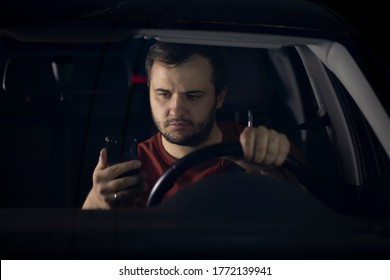 Unshaven Confident Serious Young Millenial Male Driver Sits At Steering Wheel Of Car At Night Tired Looking At Smartphone Worried Concerned About Problems. Safe Driving Concept. Loneliness Concept.