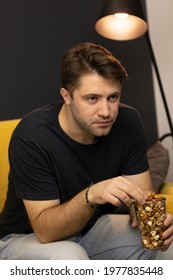 Unshaven Handsome Guy Sitting With Popcorn On Comfy Yellow Sofa Under Spot Lamp Clicks Remote Control Watching Movie On TV Or Youtube Videos. Boring Evening At Home Alone, Lockdown Copy Space Concept