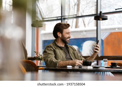 Unshaven happy man in earphones using mobile phone while drinking coffee at cafe - Powered by Shutterstock