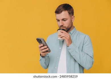 Unshaven man holding smartphone in one hand and takeaway coffee cup in another, looking at screen of phone, isolated on yellow background - Powered by Shutterstock