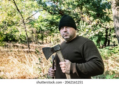 Unshaven Man Holding Two Axes In His Hands Among The Trees.