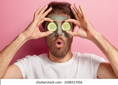 Unshaven man keeps face in good condition, covers eyes with two slices of cucumber, has clay mask on skin, cares about beauty, has rejuvenation treatments at home, stands indoor in bathroom.