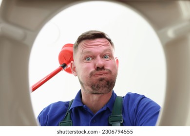 Unshaven man looks into toilet bowl with worried expression on face. Plumber prepares to clean clogged sewer pipe using red toilet plunger