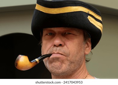 An unshaven man with a pipe in his mouth and wearing a pirate's tregoole hat.
