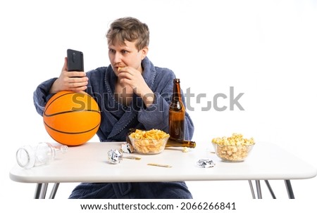 an unshaven man in a terry dressing gown sits at a table with beer, chips and watches the game, the Olympics, sports, movies, news. white, isolated background