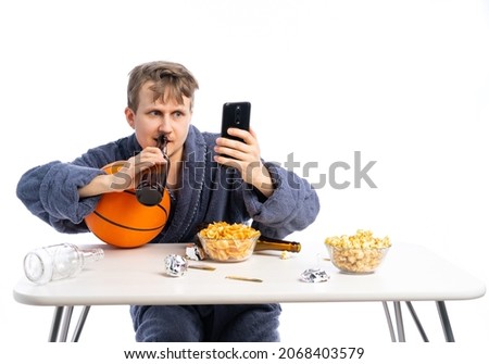 an unshaven man in a terry dressing gown sits at a table with beer, chips and watches the game, the Olympics, sports, movies, news. white, isolated background