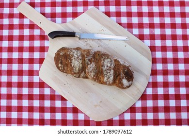 Unsliced Loaf Of Fresh Homemade Wheat Bread On A Pizza Peel And A Red And White Tablecloth.