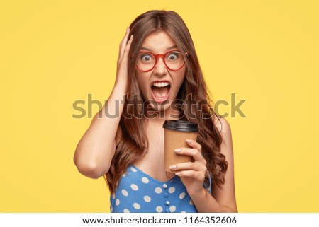 Unsound angry student shouts at her boyfriend during coffee break, holds paper disposable cup with beverage, keeps jaw dropped, exclaims in irritation, dressed in polka dot blouse. Negative feeling