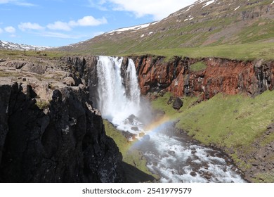 unspoiled landscape of the Icelandic east fjords with an impressive waterfall cascading down rocky cliffs and a rainbow in the spray - Powered by Shutterstock