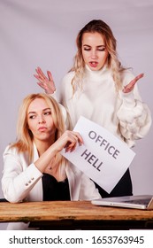 Unstable Female Boss Aggressive Gesturing And Screaming At Her Employee. Young Employee Holding Sign Office Hell. Selective Focus.