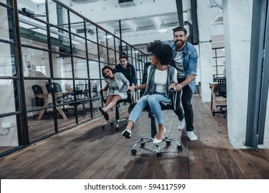 Unstoppable team! Four young cheerful business people in smart casual wear having fun while racing on office chairs and smiling