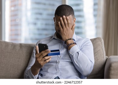 Unsuccessful payment, internet scam, fraud victim. Upset African guy sit on sofa holds card and smartphone, cover face with palm feels shocked, overspending money, lost savings, money stolen from bank - Shutterstock ID 2246712357