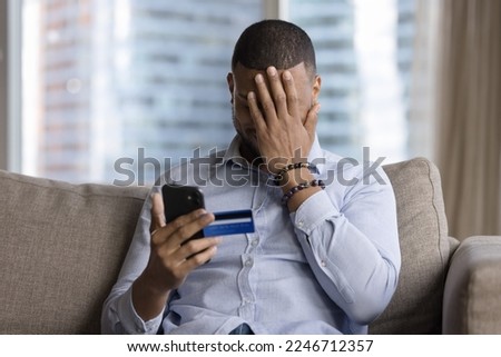 Unsuccessful payment, internet scam, fraud victim. Upset African guy sit on sofa holds card and smartphone, cover face with palm feels shocked, overspending money, lost savings, money stolen from bank
