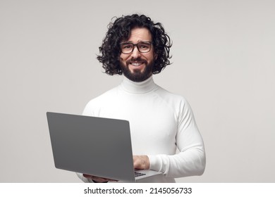 Unsure Curly Haired Male Worker Typing On Modern Netbook And Looking At Camera, With Awkward Smile With Expression Of Apologize In Studio On White Background