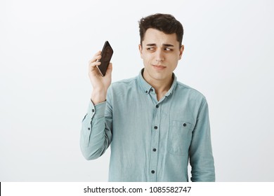 Unsure Handsome And Funny Guy With Short Dark Hair, Holding Smartphone Away Form Ear And Staring Unsure At Device, Listening Mom Yelling At Him, Standing Doubtful And Awkward Over Gray Wall