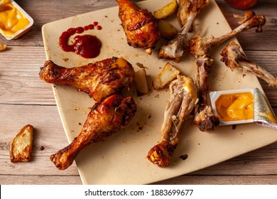 An Untidy Wooden Table With A Plate Of Deep Fried Or Grilled Seasoned Chicken Drumsticks With Half Eaten, Leftover Bones, Dipping Sauces In Packs, And Potato Chunks In Background. Food Waste Concept. 