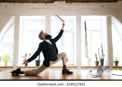 Unusual Business Process. Young Bearded Man, Office Clerk Having Fun, Doing Yoga On Wooden Table In Modern Office At Work Time With Gadgets. Concept Of Business, Healthy Lifestyle, Sport, Hobby