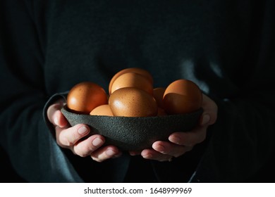 Unusual Easter on dark background. Bowl of brown eggs with hands. Darkness, rays of sunlight shine on the eggs. Concept of new life, rebirth. Rustic style. Side view, copy space