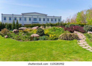 An Unusual Rectangular Building Against The Background Of A Green Flowerbed With Decorative Shrubs And Stones