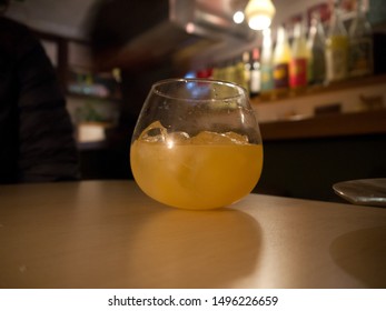 Unusual Spherical Shape Stemless Cocktail Glass With Ice Cubes And Yellow Drink On The Table, Defocused Blurred Bar Stand Background With Bottles On Shelves, Balance Concept