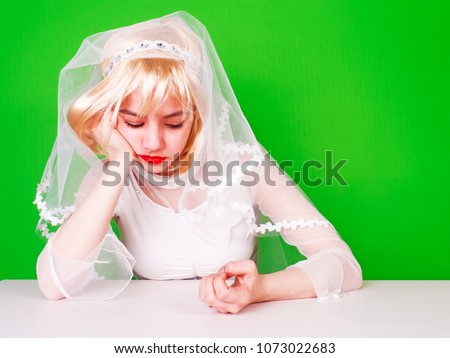 Unwanted marriage, bad life decisions concept. Serioos bride wearing white dress sitting at table and waiting for wedding, isolated on green.