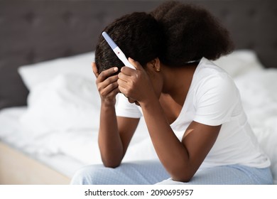 Unwanted Pregnancy Concept. Desperate African American Millennial Lady In Pajamas Sitting Alone On Bed At Home, Holding Positive Pregnancy Test And Touching Her Head, Crying, Closeup Shot, Copy Space