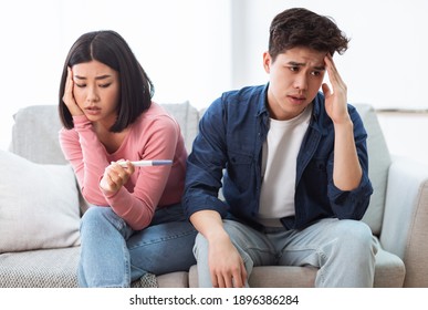 Unwanted Pregnancy. Upset Child-Free Chinese Couple Holding Positive Pregnancy Test Sitting On Sofa Indoors. Unplanned Childbirth, Unintented Pregnancies And Unhappy Parents-To-Be Concept