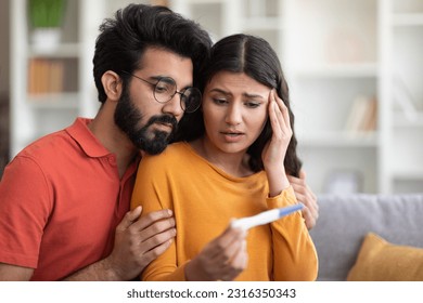 Unwanted Pregnancy. Worried young indian couple looking at positive test result, stressed millennial eastern man and woman sitting on couch at home, not ready to be parents, copy space - Powered by Shutterstock