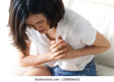 Unwell Condition And Sickness Concept; Asian Woman Having Heart Attack Sitting On Sofa. She Puts Both Hand On Her Left Chest Feeling Painful And Need Emergency Medical Assistance.