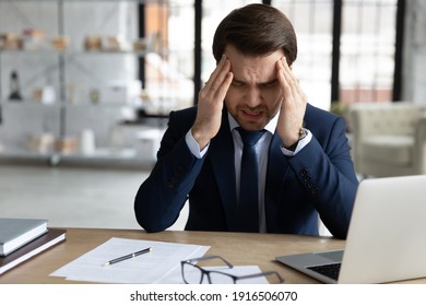 Unwell Exhausted Caucasian Young Businessman Sit In Office Working On Computer Massage Temples Suffer From Headache. Tired Unhealthy Male CEO Struggle With Migraine, Overwhelmed With Screen Job.