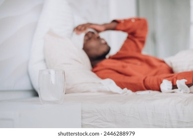 Unwell Man Lying in Bed with Water and Tissues, Concept of Sickness at Home. African American man lies in bed with his arm over his forehead, a glass of water in the foreground, capturing a moment of - Powered by Shutterstock