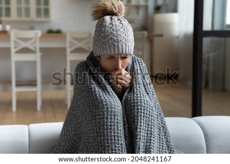 Unwell millennial female renter in hat and blanket sit in cold living room suffer from air conditioner lack. Unhealthy young Caucasian woman struggle from chill freeze at home. No heating concept.