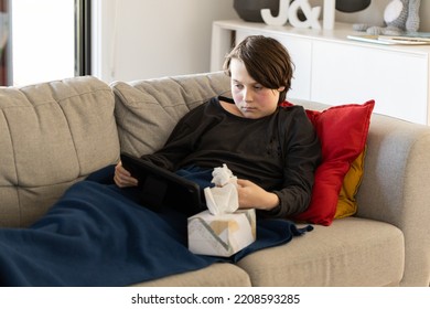 An Unwell Sick Preteen Boy Lying On A Couch Looking At His Tablet, His Cheeks Are Red From A Fever And He Has A Tissue Box Next To Him