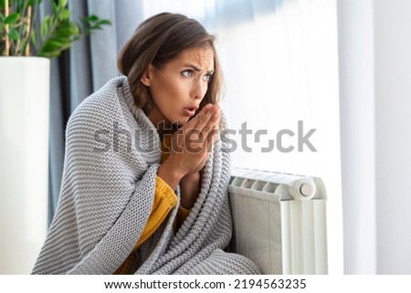 Unwell woman renter in blanket sit in cold living room hand on old radiator.suffer from lack of heat . Unhealthy young woman struggle from chill freeze at home. No heating concept.