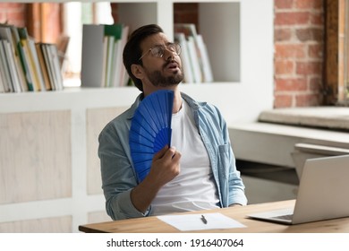 Unwell Young Caucasian Male Employee Sit On Desk Work On Computer Wave With Hand Fan. Overheated Man Worker Use Waver Suffer From Heatstroke In Office, Struggle With No AC At Workplace.