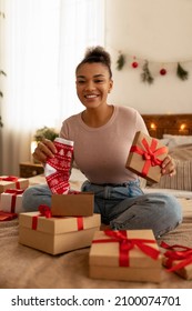 Unwrapping Gift Boxes. Excited African American Lady Receiving Christmas Present And Holding Xmas Socks, Sitting On Bed And Smiling At Camera