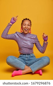 Upbeat Cheerful Dark Skinned Lady Sits With Crossed Legs On Floor, Raises Arms, Wants To Dance, Winks Eye And Smiles, Has Carefree Happy Mood, Has Rest And Fun, Makes Photo In Yellow Studio.