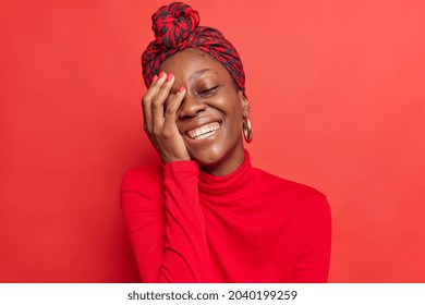 Upbeat dark skinned model keeps hand on face smiles broadly shows white teeth laughs at something expresses positive emotions wears casual clothes in one tone with background. Red color prevails