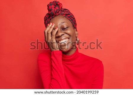 Upbeat dark skinned model keeps hand on face smiles broadly shows white teeth laughs at something expresses positive emotions wears casual clothes in one tone with background. Red color prevails