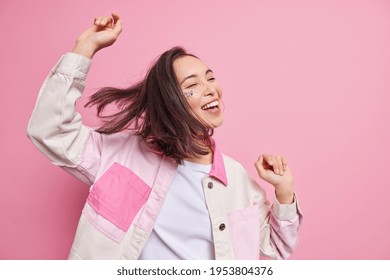 Upbeat happy brunette teenage girl being full of energy shakes hands dances carefre poses half turned against pink background celebrates victory expresses positive emotions dressed casually.