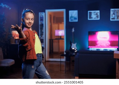 Upbeat kid records dance tutorial video in dimly lit studio for her online audience. Adorable child enthusiastically performing dancing routine on social media, participating in viral challenge