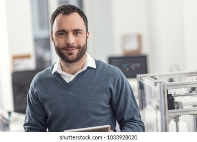 Upbeat Worker. The Portrait Of A Joyful Young Engineer Standing Near A 3D Printer In His Office, Holding A Tablet And Posing For The Camera, Smiling Pleasantly