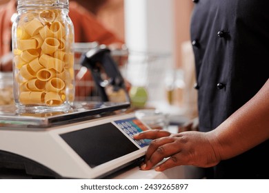 Up-close image capturing person using measuring scale, checking weight of container filled with organic bio goods. Black individual weighing glass jar of pasta at the checkout counter. - Powered by Shutterstock